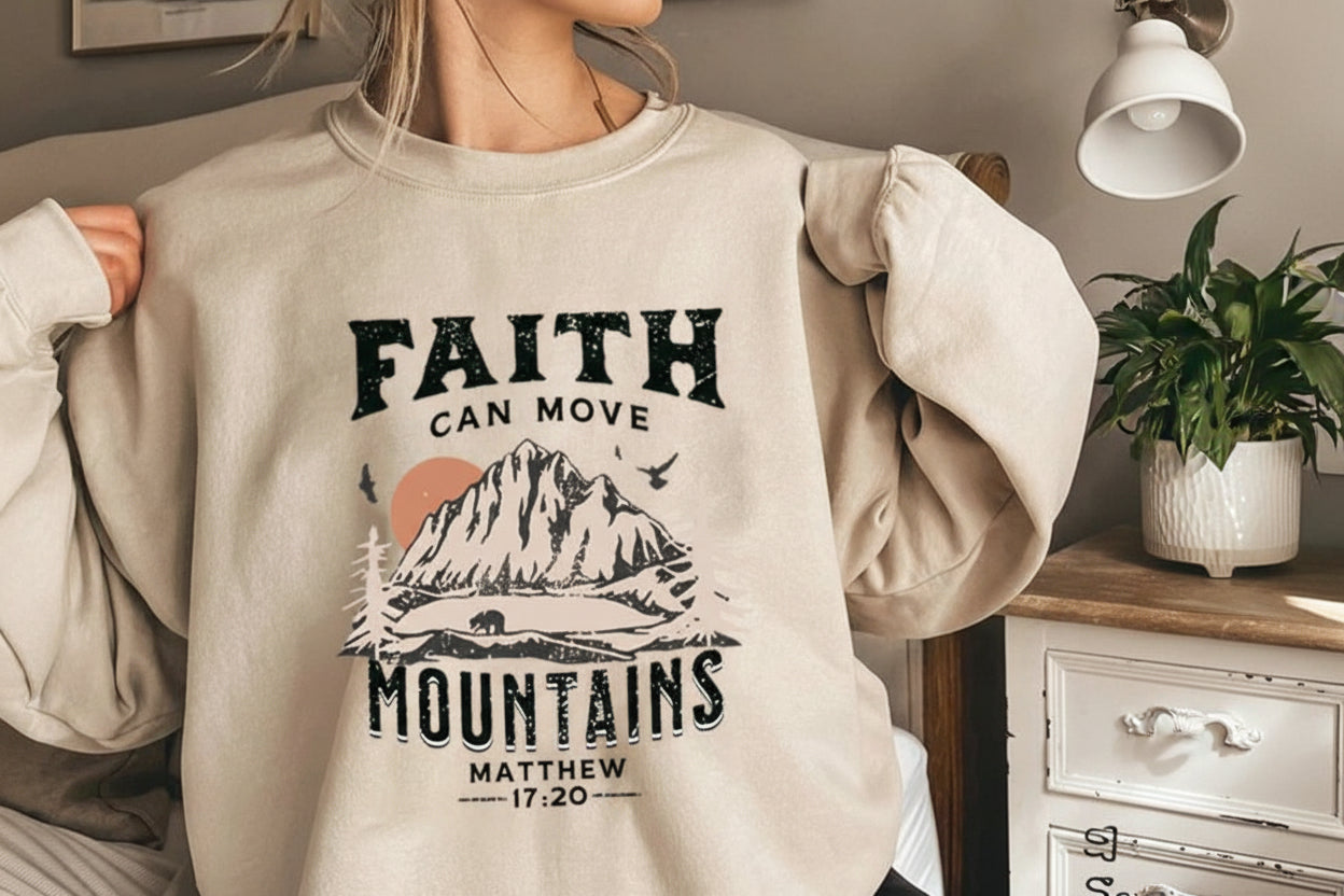 Person wearing a beige sweatshirt with a mountain graphic and 'Faith can move mountains' text.